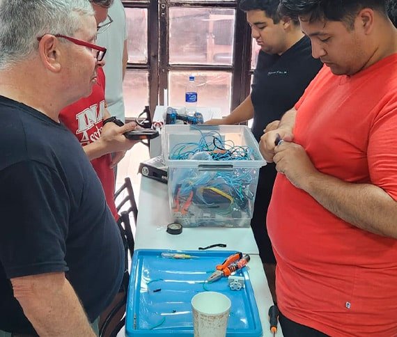 Grupo de personas trabajando alrededor de una mesa con herramientas electrónicas, alicates, cables y componentes, sugiriendo un ambiente de ensamblaje o reparación técnica.