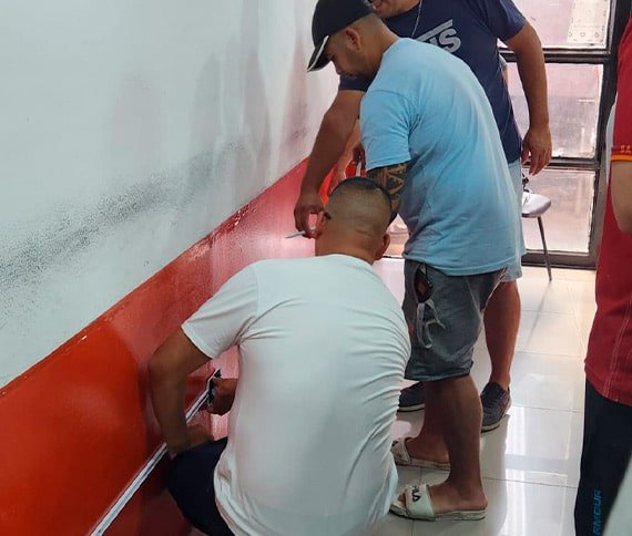 Tres personas trabajando en una pared, una arrodillada aplicando algo en la sección baja de color rojo, mientras las otras dos observan o asisten de pie. La parte superior de la pared es blanca.
