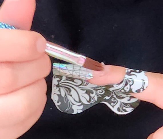 Primer plano de una mano aplicando un producto con un pincel sobre un molde decorativo de uñas en blanco y negro, usado para extensiones.