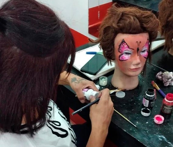 Persona trabajando en la cabeza de un maniquí, aplicando maquillaje o pintura con un pincel, rodeada de herramientas y materiales en una mesa.