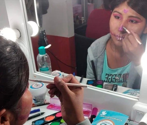 Persona aplicando maquillaje frente a un espejo iluminado, con herramientas de maquillaje visibles sobre la mesa.