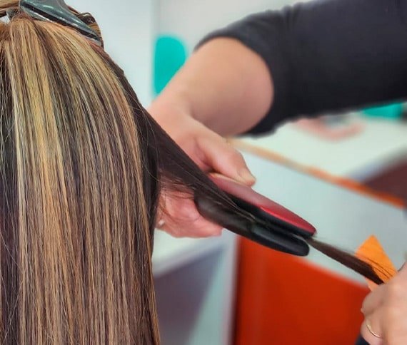 La imagen muestra un primer plano del cabello de una persona mientras se le aplica un proceso de alisado. La persona tiene el cabello largo con reflejos rubios, y otra persona está utilizando una plancha de pelo de colores rojo y negro para alisar una sección.