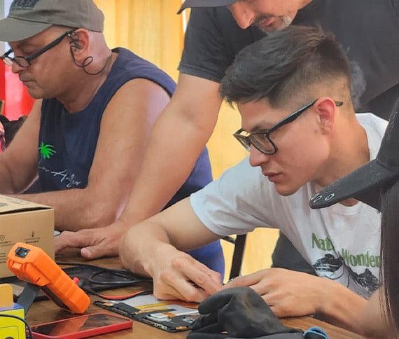 Personas trabajando en un proyecto de electrónica, utilizando herramientas como pistola de calor, soporte para circuitos, soldador y cinta adhesiva en una mesa de trabajo.