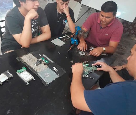 Grupo de personas trabajando en la reparación de componentes electrónicos sobre una mesa. Entre los elementos visibles hay piezas de computadora, como un disco duro y un teclado, además de herramientas como un destornillador eléctrico azul.