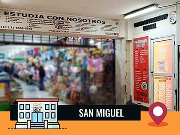 Entrada de la Sucursal de San Miguel con un cartel encima que dice: 'ESTUDIA CON NOSOTROS'. A la derecha de la entrada hay dos carteles: uno rojo que enumera servicios y otro naranja que contiene los horarios. Al final de la imagen se identifica la ubicación como 'SAN MIGUEL'.
